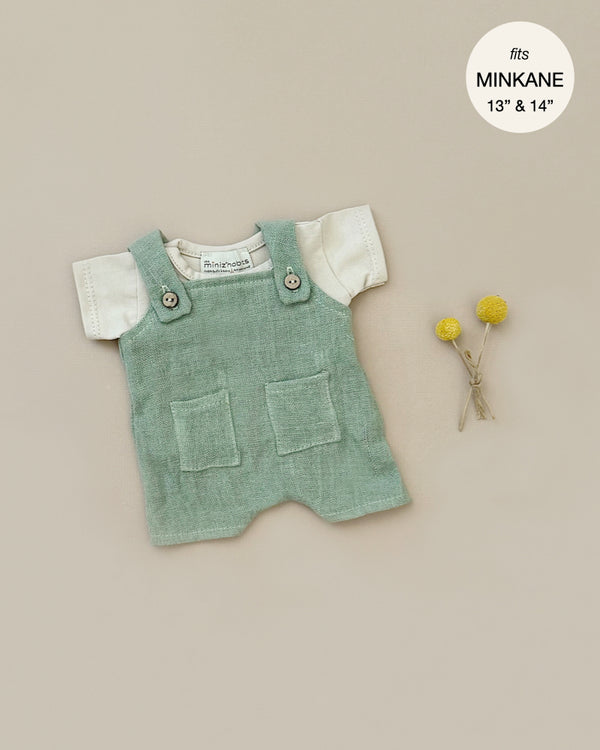 The Minikane Doll Clothing set, featuring Antonin Sage Green Linen Overalls and a Linen Jersey T-Shirt, is showcased on a beige backdrop next to two yellow billy balls. A small white tag in the upper right corner indicates that it fits Minikane Gordis dolls of 13" & 14".