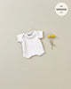 A small checkered Minikane Clothing | Honeycomb Knit Shorty Bodysuit is displayed against a beige background next to two yellow billy button flowers. Text in the top right corner reads "fits MINKANE 11", indicating it's perfect for an 11" doll accessory.