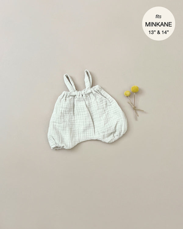 A small, light-colored romper made of double gauze with straps is displayed on a plain beige background. Two yellow flowers with stems are placed to the right of the romper. A white circular label in the top right corner reads "fits Minikane Gordis dolls 13’’ & 14’’.” The product name is Minikane Doll Clothing | Kim Bloomers in Raw Cotton.