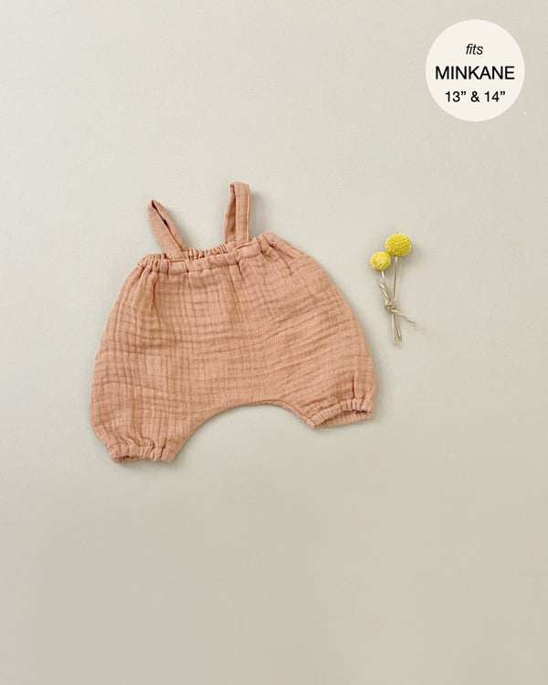 A pair of light brown baby overalls made from double gauze fabric with shoulder straps laid flat on a beige background next to two small yellow flowers. The text "fits MINKANE 13'' & 14''" is displayed inside a white circle in the top-right corner, perfect for Minikane Doll Clothing | Kim Bloomers in Marsala.