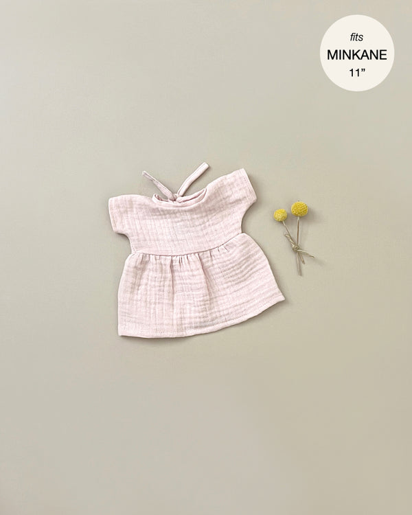 A small, light pink Minikane Doll Clothing | Faustine Dress and Bonnet in Petal Pink with short sleeves and a bow at the neckline is displayed on a neutral background. Next to the dress are two small yellow flowers. A circular label in the top right corner reads "fits Minikane Babies 28cm dolls.