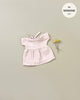 A small, light pink Minikane Doll Clothing | Faustine Dress and Bonnet in Petal Pink with short sleeves and a bow at the neckline is displayed on a neutral background. Next to the dress are two small yellow flowers. A circular label in the top right corner reads "fits Minikane Babies 28cm dolls.