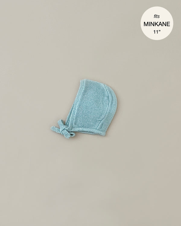 A small, light blue knitted bonnet with a chin tie is laid flat against a plain beige background. There is a circular label in the top right corner that reads "Minikane Doll Clothing | Élie Blue Knit Bonnet".