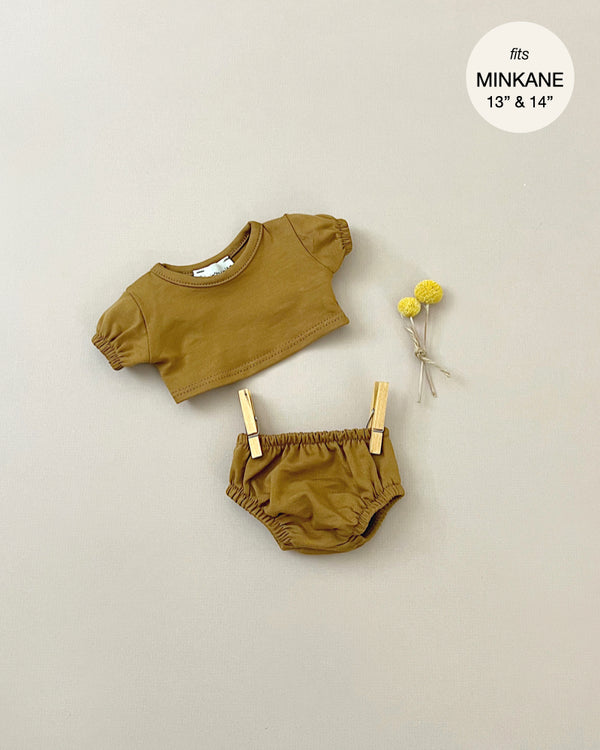 Tan-colored baby outfit, featuring a balloon-sleeved t-shirt and Charlotte jersey panties, displayed against a beige background. Two small yellow flowers and a pair of clothespins are placed nearby. A circular label in the top right corner says "Minikane Clothing | Charlotte Balloon Sleeve Shirt and Briefs fits Minikane Gordis dolls 13" & 14".