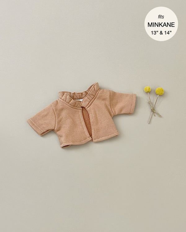A small, light brown Minikane Doll Clothing | Claudia Cardigan in Brown Sugar Fleece Doll Clothing with a ruffled neckline is displayed on a beige background. The text in the top right corner reads "fits MINKANE 13” & 14”." Two small yellow flowers are placed to the right of the cotton jacket.