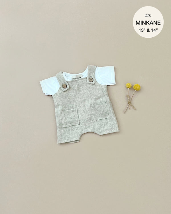 A white jersey t-shirt set with a beige textured romper featuring front pockets and buttoned shoulder straps is laid out on a plain background. Two small yellow pom-pom flowers are placed beside the outfit. There is a circular label stating "fits Minikane Gordis dolls 13" & 14'. The product shown is the Minikane Doll Clothing | Antonin Overalls Set Doll Clothing.