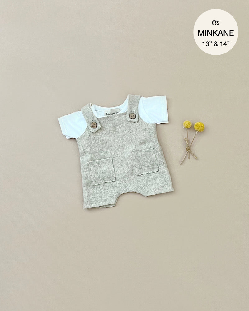 A white jersey t-shirt set with a beige textured romper featuring front pockets and buttoned shoulder straps is laid out on a plain background. Two small yellow pom-pom flowers are placed beside the outfit. There is a circular label stating "fits Minikane Gordis dolls 13" & 14'. The product shown is the Minikane Doll Clothing | Antonin Overalls Set Doll Clothing.