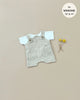 A white jersey t-shirt set with a beige textured romper featuring front pockets and buttoned shoulder straps is laid out on a plain background. Two small yellow pom-pom flowers are placed beside the outfit. There is a circular label stating "fits Minikane Gordis dolls 13" & 14'. The product shown is the Minikane Doll Clothing | Antonin Overalls Set Doll Clothing.