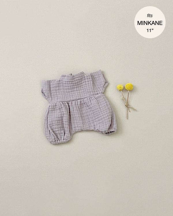 A small, light purple, textured Minikane Clothing | Romper and Bonnet Set with short sleeves is displayed on a beige background. To the right of the romper, there are two yellow billy ball flowers. In the top right corner, there's a text overlay reading "fits Minikane Babies 11".