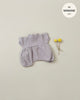 A small, light purple, textured Minikane Clothing | Romper and Bonnet Set with short sleeves is displayed on a beige background. To the right of the romper, there are two yellow billy ball flowers. In the top right corner, there's a text overlay reading "fits Minikane Babies 11".