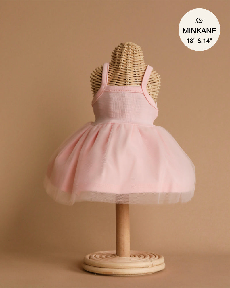 The Minikane Doll Clothing Rosella Tutu in Pale Pink, a sleeveless, tutu-style dress designed for 13" and 14" Minikane dolls, is beautifully displayed on a wooden mannequin stand against a neutral beige background. This hand washable dress features a fitted bodice with a voluminous tulle skirt and is elegantly designed in France.