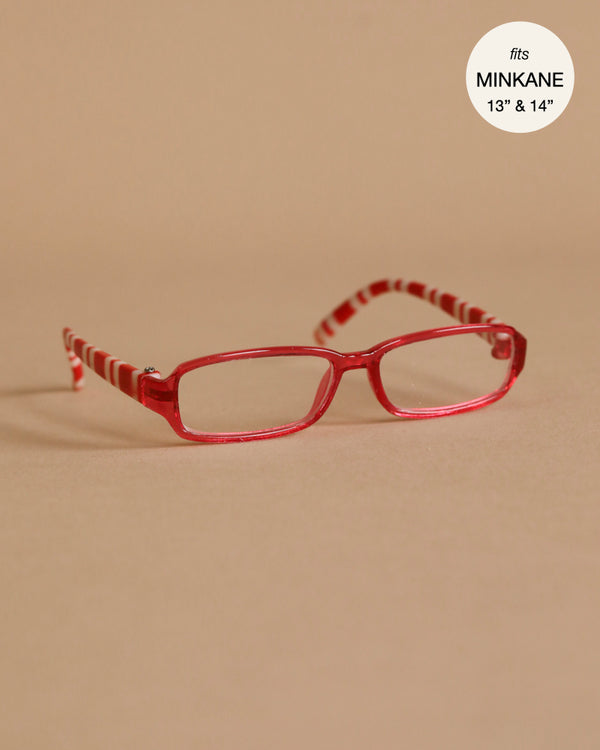 A pair of red and white striped glasses, perfect as Minikane dolls accessories, is displayed on a flat, light brown surface. The text "fits MINKANE 13\" & 14\"" is written in a white circle at the top right corner. These stylish glasses are part of the Minikane Doll Clothing collection and are specifically named TESSA Doll Glasses.