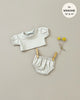 A light grey doll outfit, part of the Les P'tits Basics collection, is displayed against a beige background. The set includes a balloon-sleeved t-shirt and elasticated shorts. Wooden clothespins clip the shorts to a string, while two yellow billy button flowers lay beside the outfit. Text reads "Minikane Doll Clothing | Charlotte Balloon Sleeve T-Shirt and Briefs Set in Linen Ecru fits Minikane Gordis dolls 13' & 14'".