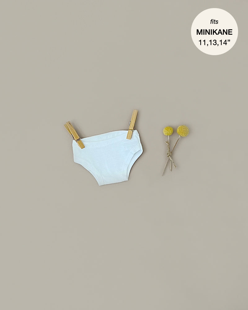 A pair of white cotton Minikane Doll Clothing | Culotte in White Cotton is clipped to a string with two wooden clothespins. To the right, there are two small yellow flowers with thin stems. In the upper right corner, a gray circle reads, "fits MINIKANE 11,13,14"." The background is a light beige color.