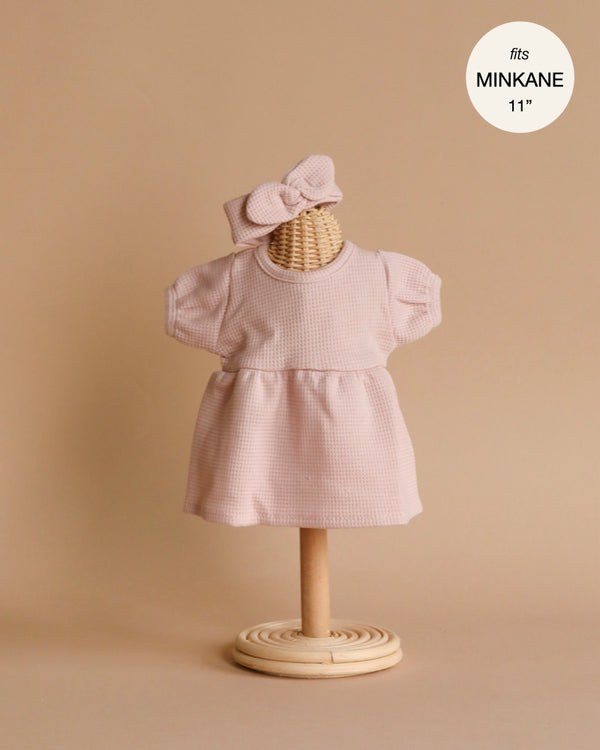 A petite Faustine dress in petal pink, complete with delicate balloon sleeves and a matching bow headband, is elegantly displayed on a minimalist wooden stand against a beige background. The text in the top right corner reads "fits MINKANE 11" or 28cm Babies dolls.