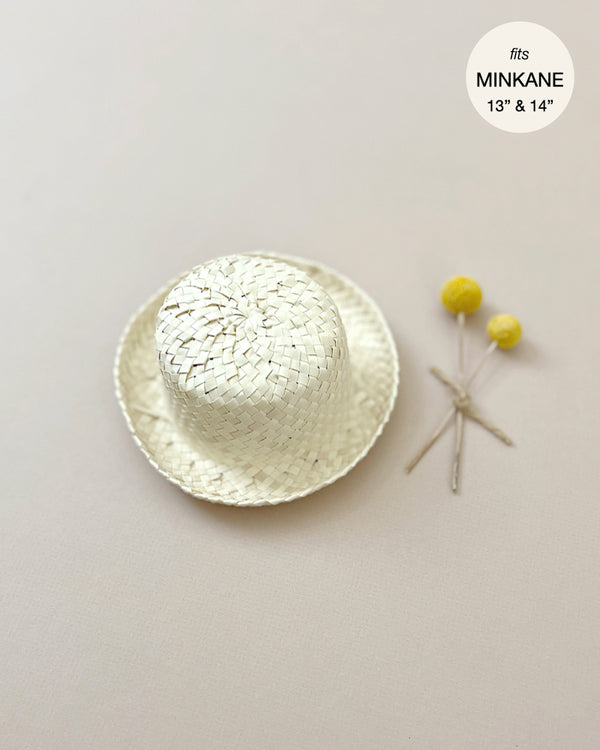 A small woven Minikane | Doll Straw Hat designed for 13" and 14" dolls by MINKANE is displayed on a light-colored surface. Designed in France, the hat is paired with two yellow pompom-like accessories tied together. A label indicates the cotton hat fits dolls of specified sizes.