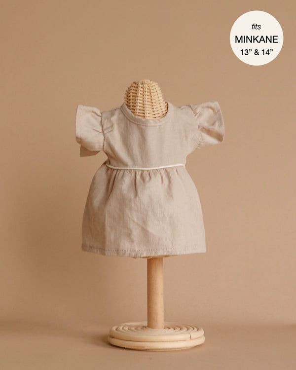 A beige, short-sleeved doll dress from the Minikane Doll Clothing collection is displayed on a small dress form with a round, textured head and a simple circular wooden base. The background is neutral. In the top right corner, text reads "fits Minikane dolls 13" & 14". This charming Daisy Dress with Headband, designed in France and hand washable, is perfect for any collection.