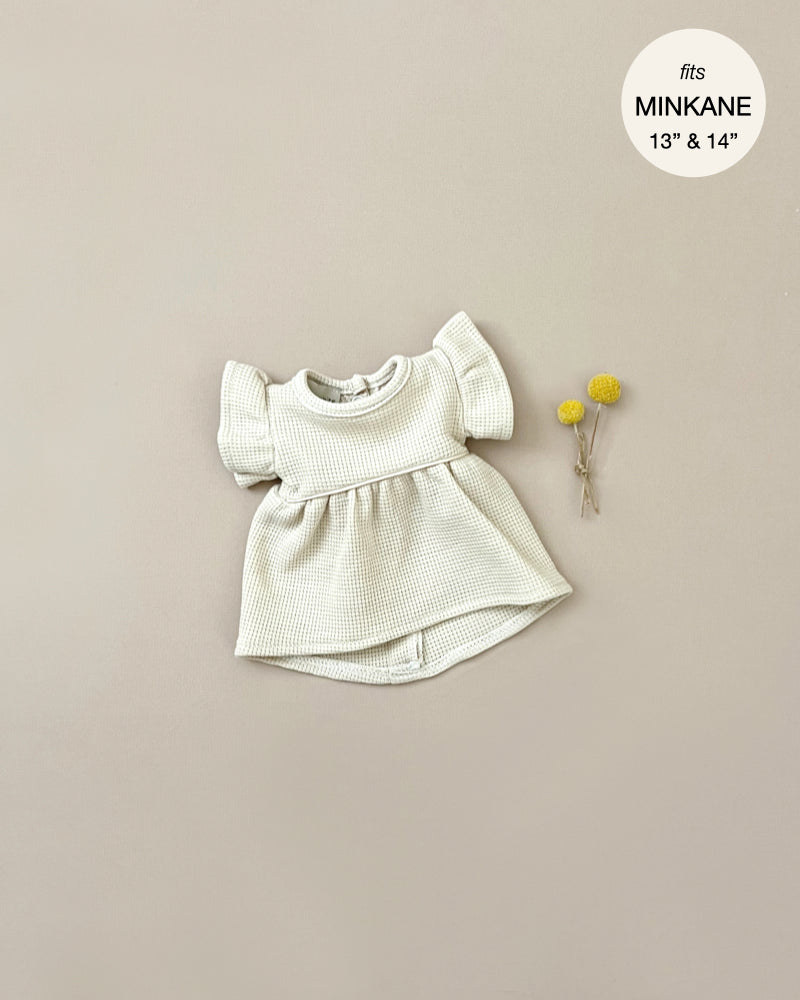 A light cream-colored, short-sleeve Minikane Doll Clothing | Linen Dress and Headband Set with a honeycomb mesh pattern is laid flat on a neutral background. To the right of the dress, there are two small yellow flowers. A circular label at the top right indicates the dress fits Minikane Gordis 13” & 14” dolls.