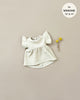 A light cream-colored, short-sleeve Minikane Doll Clothing | Linen Dress and Headband Set with a honeycomb mesh pattern is laid flat on a neutral background. To the right of the dress, there are two small yellow flowers. A circular label at the top right indicates the dress fits Minikane Gordis 13” & 14” dolls.