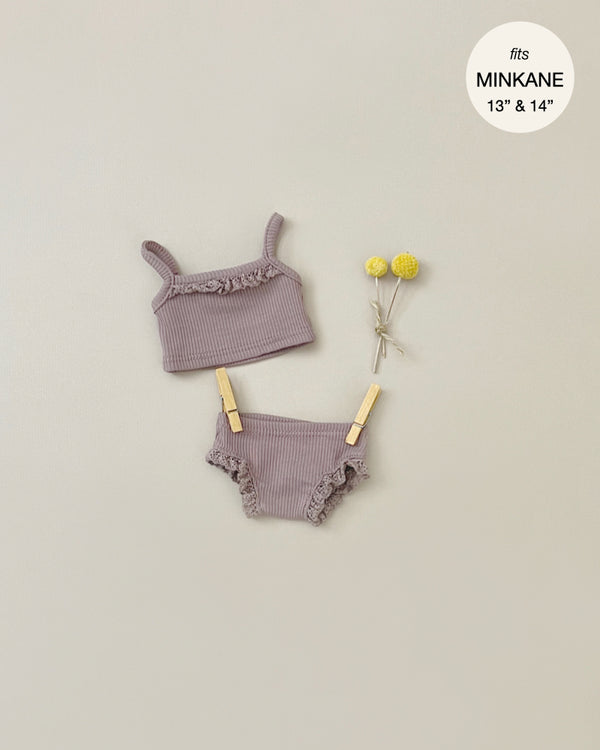 A two-piece, mauve ribbed knit and lace doll outfit with a sleeveless top and matching bottom, is pinned to a neutral background beside three yellow ball-like flowers. The text indicates it fits 13" and 14" Minikane Gordis dolls. This product is the **Minikane Doll Clothing | Underwear in Dark Orchid Ribbed Knit**.