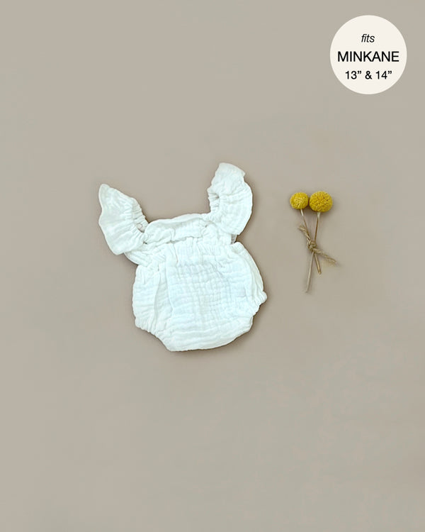 A white, ruffled baby garment in soft double gauze is laid flat on a light beige surface. Two small yellow flowers, tied together, are placed beside it. Text in the corner reads "Minikane Doll Clothing | Lou Romper in Cream Cotton - fits Minikane Gordis dolls 13" & 14".