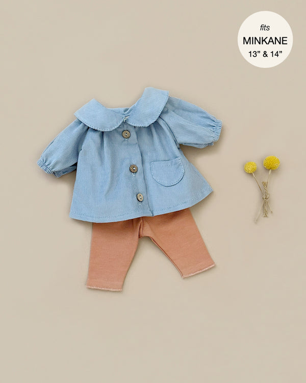 The Minikane Doll Clothing set includes a light blue button-up blouse featuring long sleeves, a Peter Pan collar, and a small pocket, paired with brown sugar jersey leggings. Yellow flowers are displayed beside the outfit. In the top right corner, there's a white circle that reads "fits Minikane Gordis dolls 13'' & 14''.