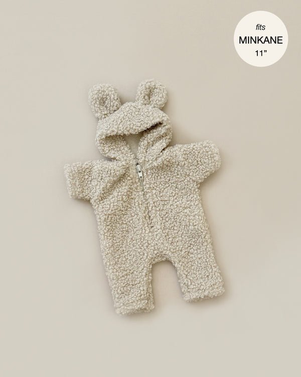 The Minikane Doll Clothing Winnie Beige Terry Jumpsuit is a beige, fluffy teddy bear-style onesie with a hood that features bear ears, designed to fit an 11-inch Minikane doll. The tag in the top right corner reads "fits MINIKANE 11"" and this cozy jumpsuit includes a front zipper for closure.