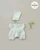 The Minikane | Noa Double Gauze and Bonnet Set in Linen Ecru Doll Clothing features a set of baby clothes in a soft double gauze fabric, displayed against a beige background. The set includes a bonnet with chin ties and a short-sleeved romper with bloomer-style legs. Nearby, two small yellow dried flowers are tied together with string. Text reads, "fits Minikane Babies.