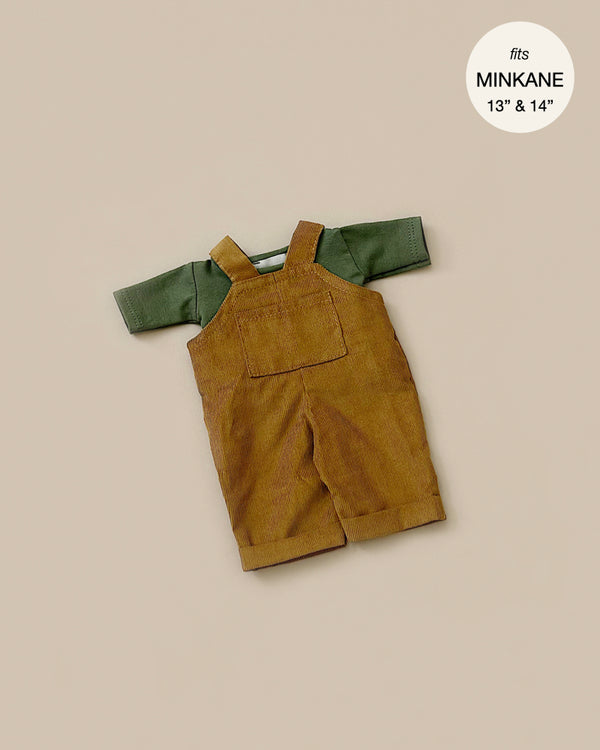 The Minikane Doll Clothing | Antoine Corduroy Overalls Set, featuring a green short-sleeve shirt paired with brown corduroy overalls, is displayed on a beige surface. The text inside a circle reads "fits Minikane Gordis dolls 13" & 14".