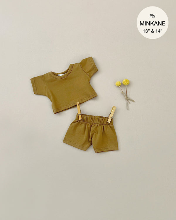 A small camel Vito t-shirt and shorts set is laid out on a beige surface. Designed for Minikane Gordis dolls, the outfit includes a short-sleeved shirt secured with tiny wooden clothespins. Yellow billy balls flowers are placed beside it. The text above reads "fits MINKANE 13" & 14".