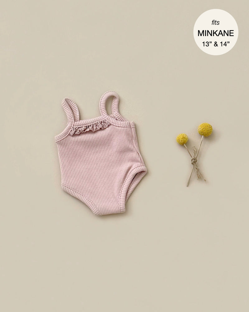 A pale pink, ribbed baby onesie with shoulder straps is displayed on a beige background. To the right of the onesie, two small yellow flower blossoms are tied together. Above, in a white circle, it states "fits Minikane Gordis dolls 13'' & 14'' dolls." This Les P'tits Basique Jumpsuit in Petal Ribbed Knit is the perfect addition to your doll clothing accessories collection.