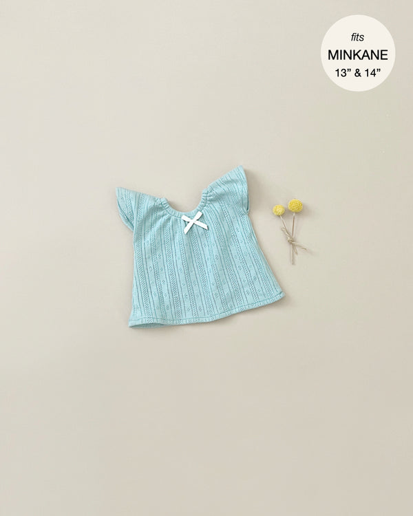 A small, blue, short-sleeve dress with subtle vertical patterns, tied with a white bow in the center of the neckline, is laid out on a beige surface. Two small yellow flowers with stems are placed to the right of the dress. The text indicates it fits Minikane Gordis dolls of 34 & 37cm. This is the Minikane Clothing | Nightgown in Peacock Blue.