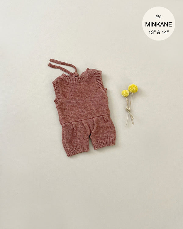 A Minikane | Orlando Heather Caramel Knit Romper Doll Clothing for a 13 to 14-inch doll, displayed on a light beige background. The sleeveless overalls, made from a hand washable knit composition, include a string for tying. Two small yellow flowers lie next to the outfit.