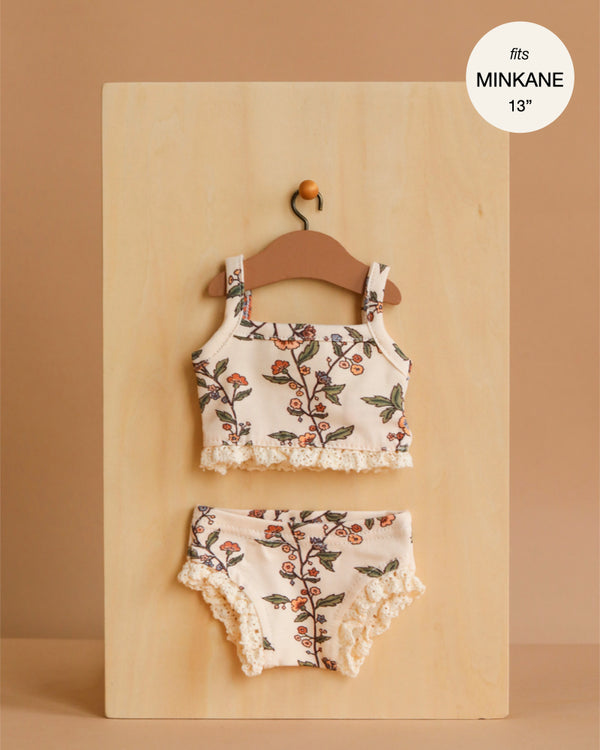 A Minikane 13" Doll Clothing | Les P'tits Basiques, 2-Piece Set in Jersey - Gisele, featuring a floral sleeveless top and ruffled bottoms, hangs on a wooden hanger. Label: fits Minikane dolls 13", made in France.