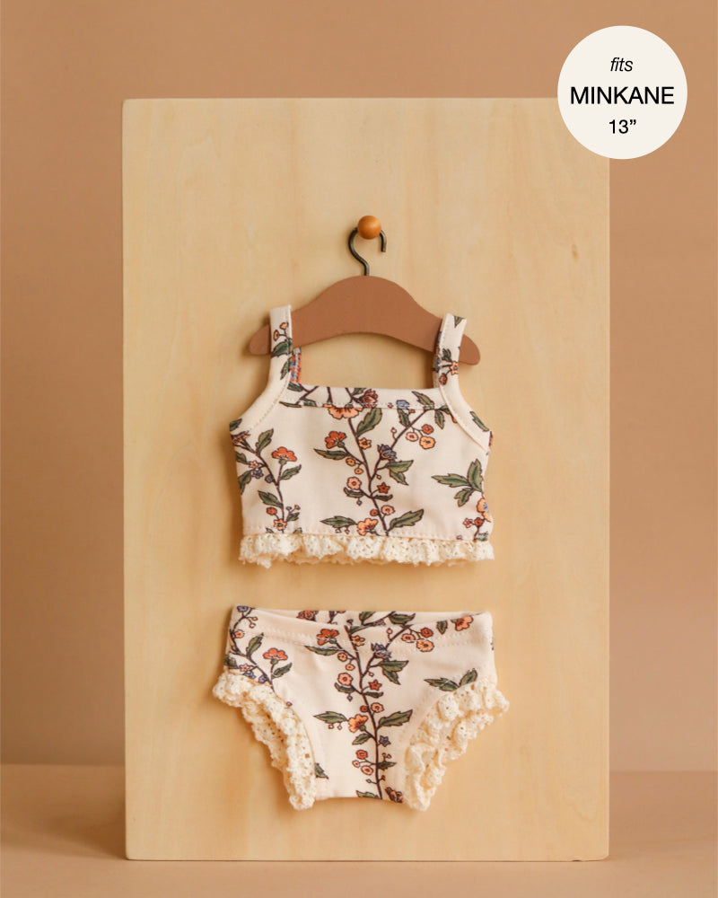 A Minikane 13" Doll Clothing | Les P'tits Basiques, 2-Piece Set in Jersey - Gisele, featuring a floral sleeveless top and ruffled bottoms, hangs on a wooden hanger. Label: fits Minikane dolls 13", made in France.