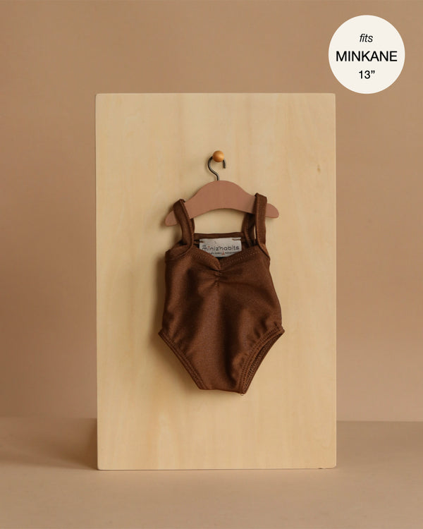 A Minikane 13" Doll Clothing Anna Leotard in Shiny Lycra - Chocolate Brown hangs on a wooden hanger against a light wood board, labeled "fits Minikane Gordis dolls 13 inch." Expertly made in France for your doll collection.