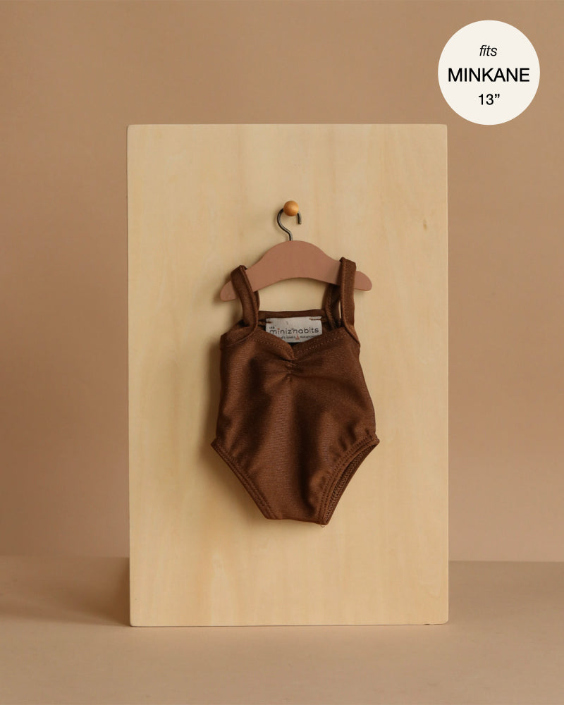 A Minikane 13" Doll Clothing Anna Leotard in Shiny Lycra - Chocolate Brown hangs on a wooden hanger against a light wood board, labeled "fits Minikane Gordis dolls 13 inch." Expertly made in France for your doll collection.