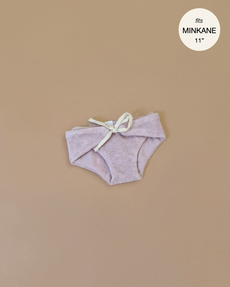 The Minikane Doll Clothing in Petal Pink is crafted as doll-sized shorts with a bow in soft double gauze cotton, displayed on a beige background, and features a circular label in the top-right corner stating "fits Minikane Babies 11".