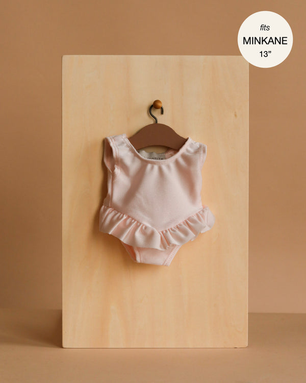 The Maia Ruffled Leotard in shiny pink lycra hangs on a wooden hanger against a light board, labeled “fits Minikane Gordis dolls” and is perfect for 13-inch dolls.