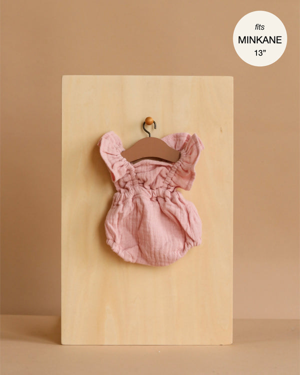 The Minikane 13" Doll Clothing | Lou Retro Romper in Cotton Gauze - Petal Pink hangs on a small wooden hanger against a light wood board, with a label stating it fits Minikane Gordis dolls 13'' on a beige background.