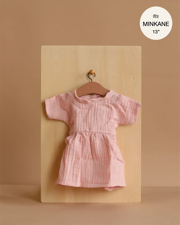 The Minikane 13" Doll Clothing | Faustine Dress in Cotton Gauze - Petal Pink, a light pink short-sleeve dress, hangs on a wooden hanger against a beige background. A label above reads, "fits Minikane Gordis dolls 13'' & 14''.
