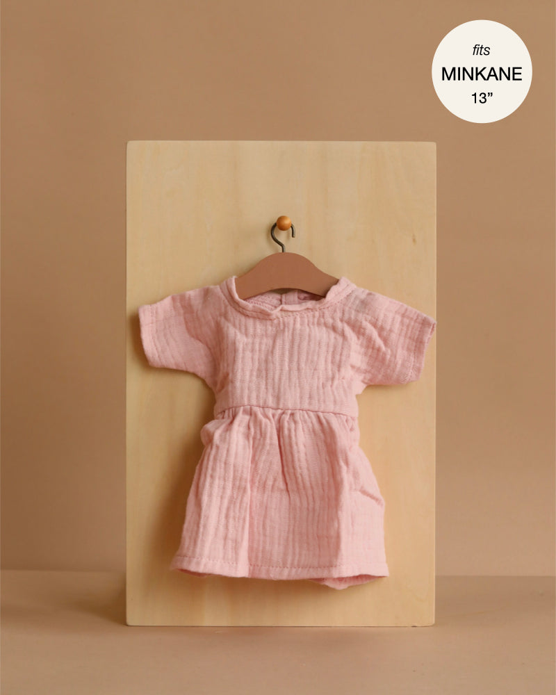 The Minikane 13" Doll Clothing | Faustine Dress in Cotton Gauze - Petal Pink, a light pink short-sleeve dress, hangs on a wooden hanger against a beige background. A label above reads, "fits Minikane Gordis dolls 13'' & 14''.