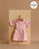 The Minikane 13" Doll Clothing | Faustine Dress in Cotton Gauze - Petal Pink, a light pink short-sleeve dress, hangs on a wooden hanger against a beige background. A label above reads, "fits Minikane Gordis dolls 13'' & 14''.