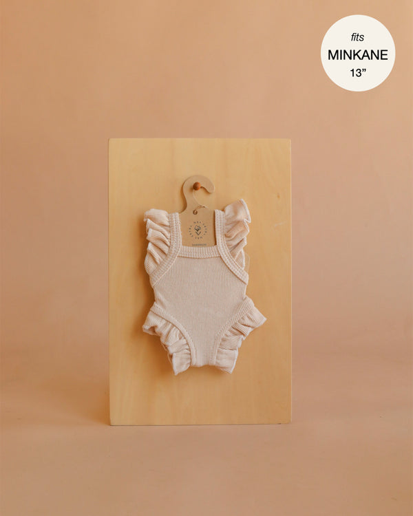 An ivory frilled bodysuit from Mrs. Ertha Doll Clothes, designed to fit 13″-14″ dolls, is displayed on a hanger against a wooden board. Crafted from organic cotton with ribbed texture and ruffled sleeves, this piece is perfect for collectors seeking stylish Minikane doll clothes. The warm beige background features text in the top right corner that states “fits MINIKANE 13′’”.