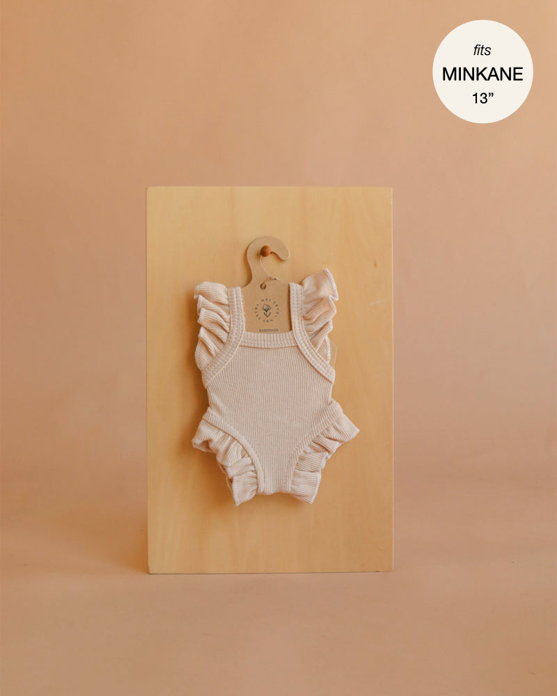 An ivory frilled bodysuit from Mrs. Ertha Doll Clothes, designed to fit 13″-14″ dolls, is displayed on a hanger against a wooden board. Crafted from organic cotton with ribbed texture and ruffled sleeves, this piece is perfect for collectors seeking stylish Minikane doll clothes. The warm beige background features text in the top right corner that states “fits MINIKANE 13′’”.