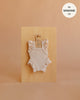 An ivory frilled bodysuit from Mrs. Ertha Doll Clothes, designed to fit 13″-14″ dolls, is displayed on a hanger against a wooden board. Crafted from organic cotton with ribbed texture and ruffled sleeves, this piece is perfect for collectors seeking stylish Minikane doll clothes. The warm beige background features text in the top right corner that states “fits MINIKANE 13′’”.