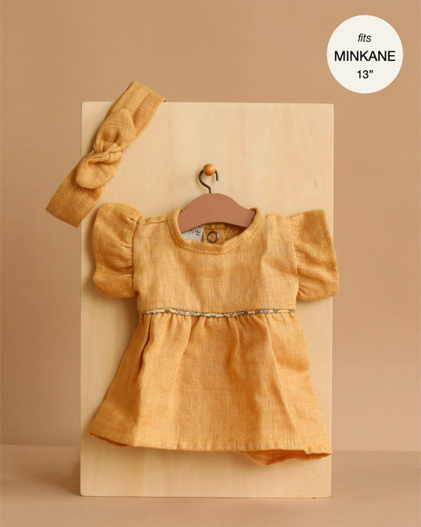 A Minikane Doll Daisy Dress & Headband in washed linen oatmeal, featuring puffed sleeves, is displayed on a wooden board with a label: "fits Minikane dolls 13''"—ideal for your 13 inch doll clothes collection.