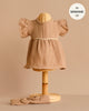 A small woven mannequin models the Minikane Doll Clothing Daisy Dress & Headband in Cotton Gauze - Latte, featuring a short sleeve ruffled dress with white trim for 13-inch dolls, perfect to enhance your 13 inch doll wardrobe.