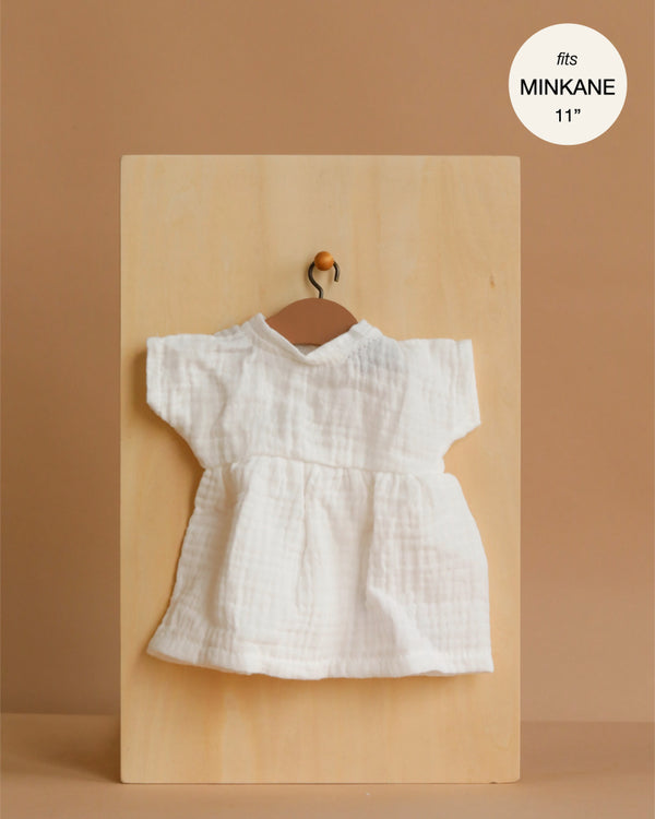 The Minikane 11" Doll Clothing | Faustine Dress in Cotton Gauze - White is a small white short-sleeved dress for 11-inch dolls, shown on a wooden hanger with a soft brown background and "fits MINIKANE 11''" label in the corner.