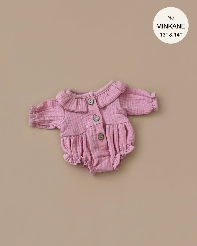 The Leonore Ruffled Jumpsuit in Tea Pink for Minikane dolls is a pink, long-sleeve button-up romper with ruffled cuffs and a round collar made of soft cotton, displayed on a beige background and fits 13" to 14" sizes.